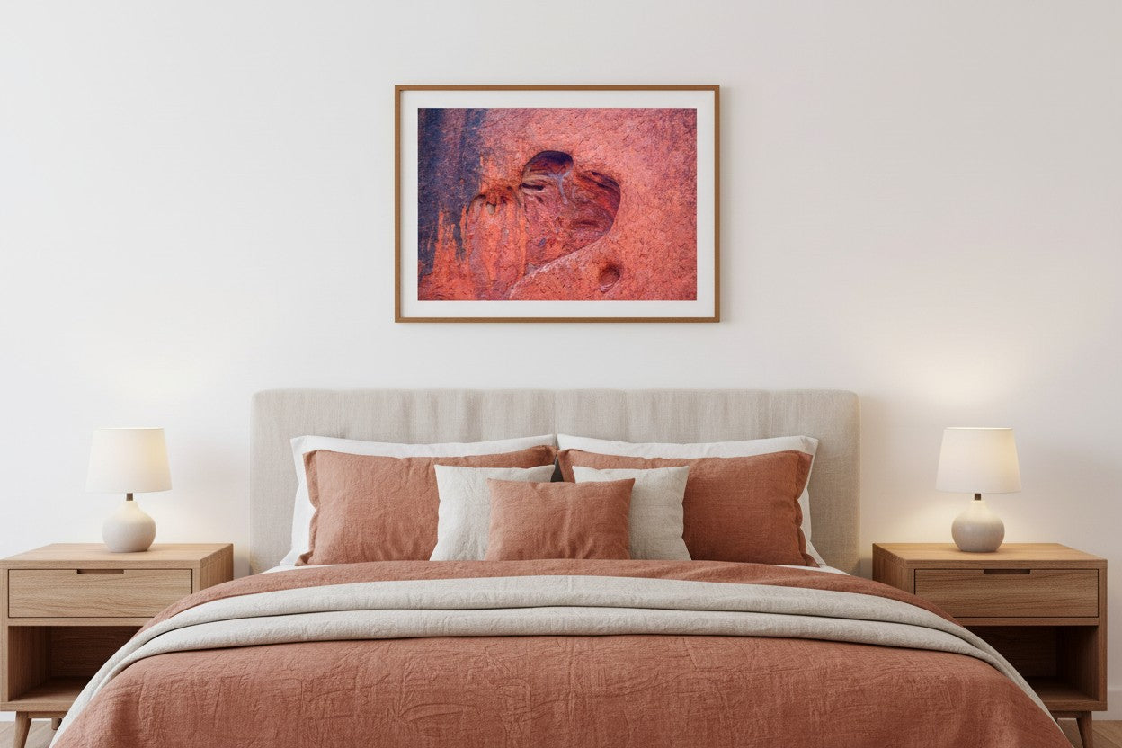 Heart of Uluṟu fine art print in a thin timber frame above a bed in a warm romantic bedroom with cream and terracotta linen — unframed Australian wall art