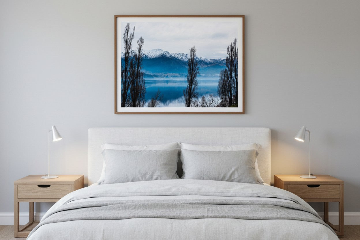 Lake Hayes fine art print in a thin timber frame above a bed in a calm cool-toned bedroom with white and grey linen — unframed New Zealand wall art
