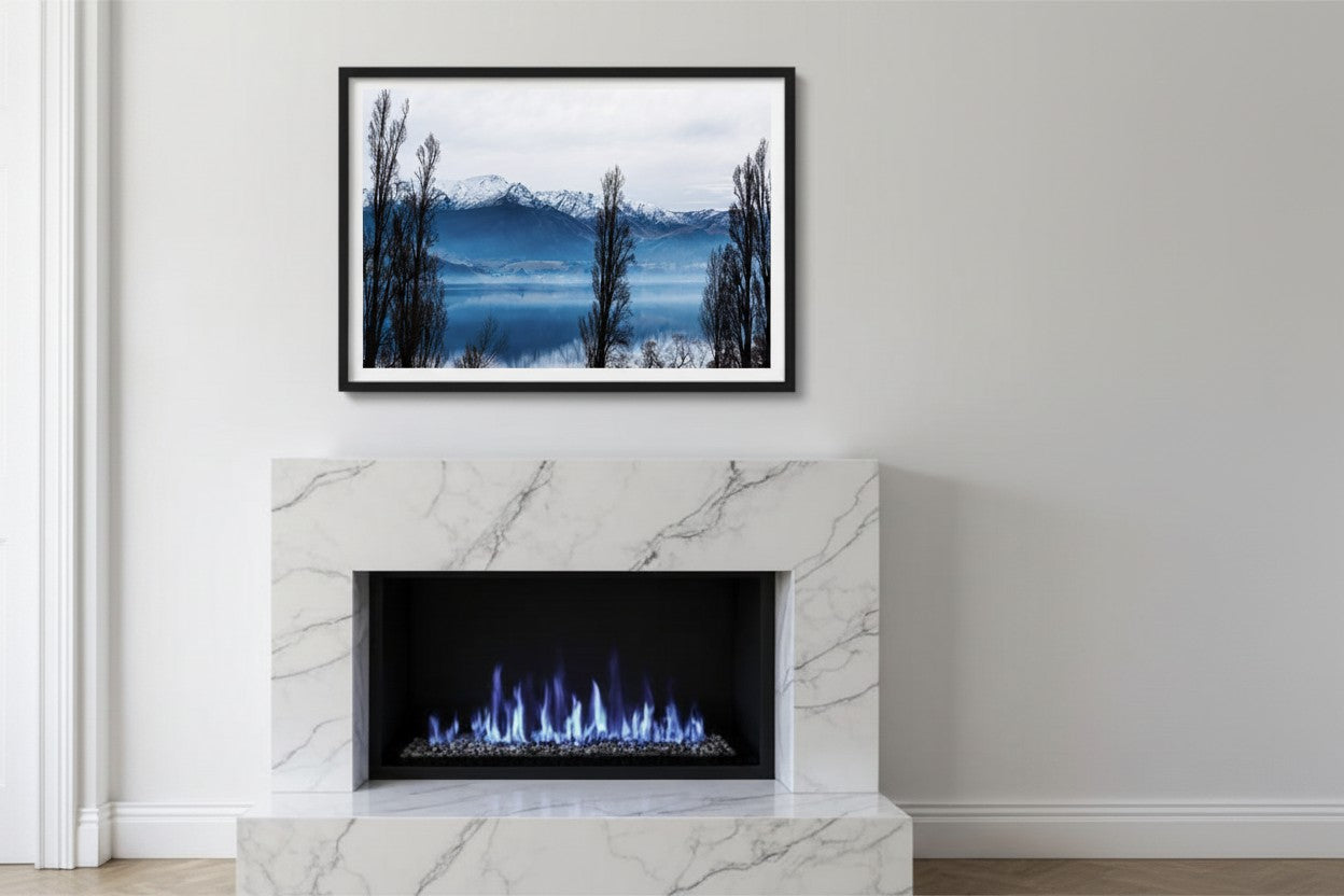 Lake Hayes fine art print in a thin black frame centered above a luxury white marble fireplace — unframed New Zealand landscape wall art