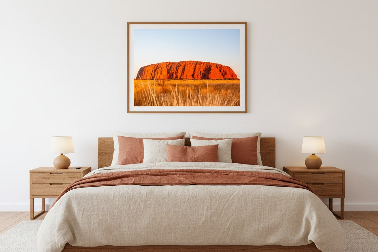 Uluṟu fine art print in a thin timber frame above a bed in a warm Australian-inspired bedroom with cream and terracotta linen — unframed landscape wall art